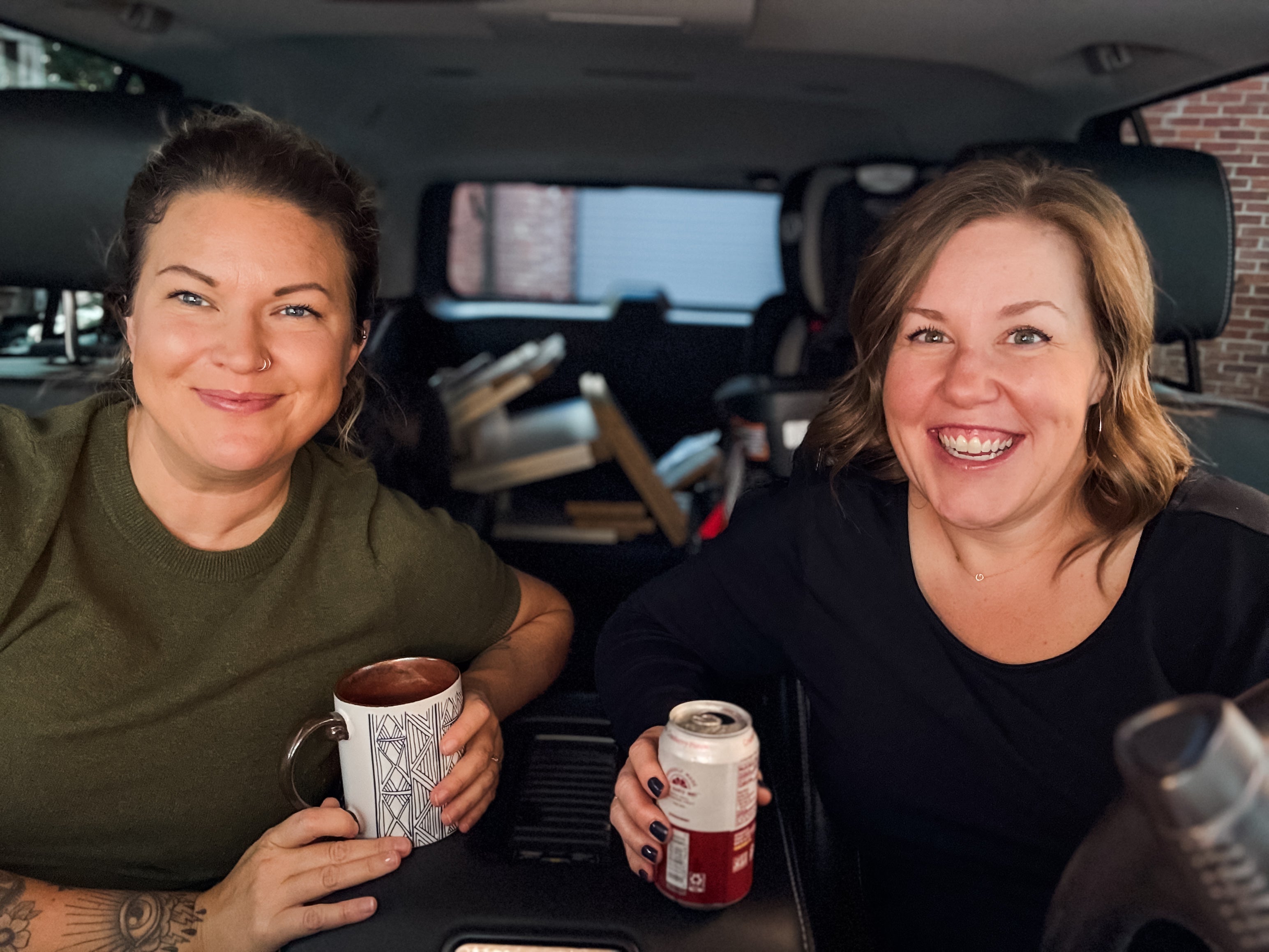 Two women sitting inside a vehicle, one holding a mug and the other a can, smiling. Don't Do It Podcast